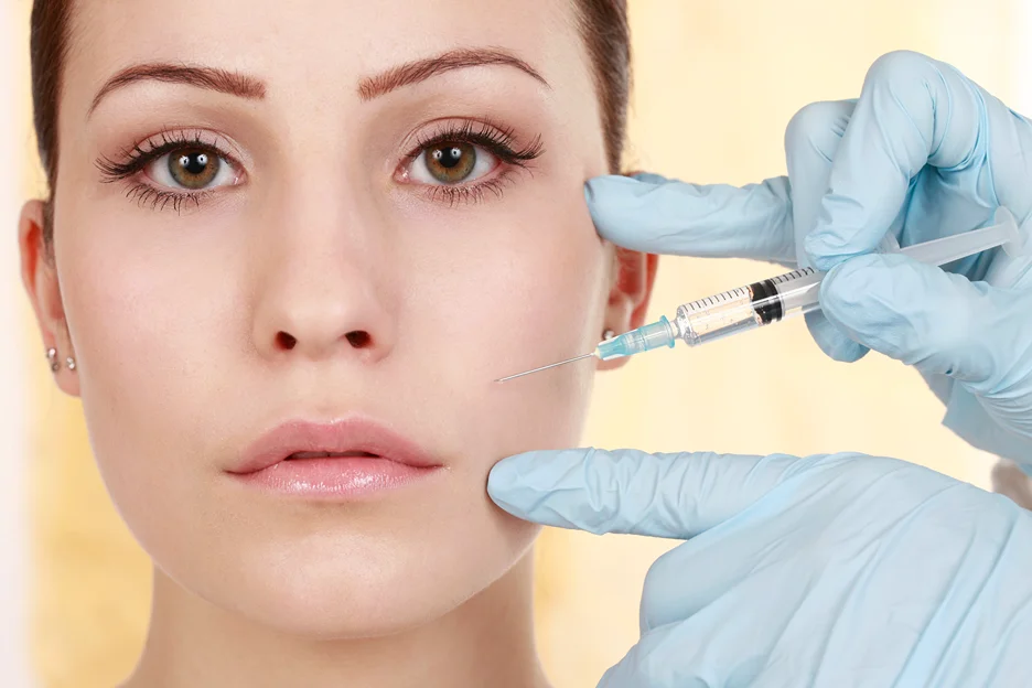 Gloved practitioner holding a syringe to a woman's cheek for filler