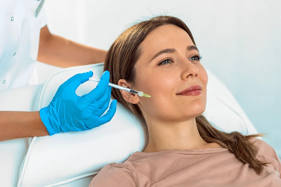 Gloved practitioner injecting cheek filler into a reclining woman
